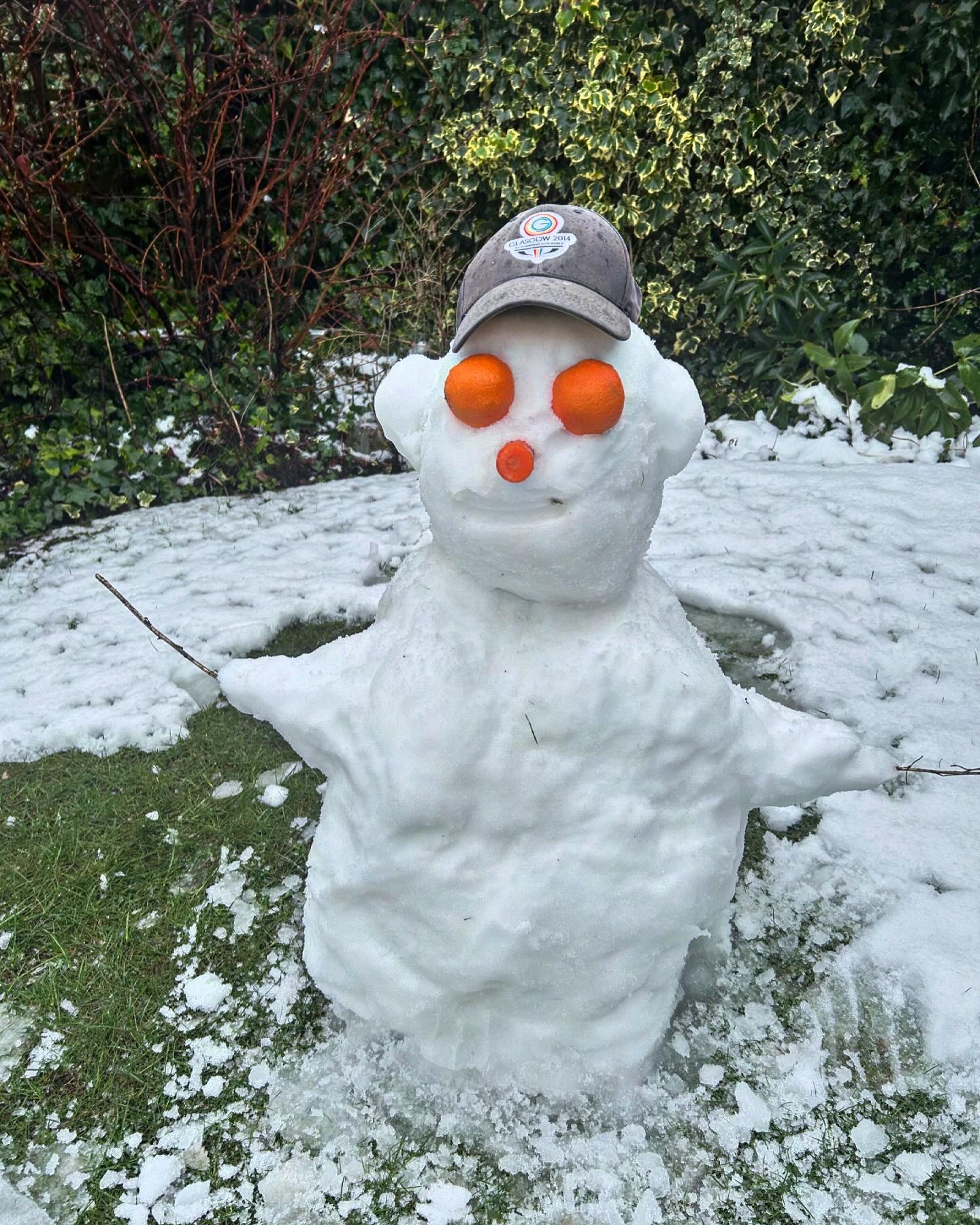 Photo of a snow person, the snow person is quite small, around 80cm, and has a carrot nose, satsuma eyes, and wears a peaked cap. It sits on the grass; CC-BY-NC